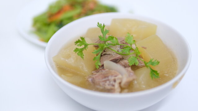  A white bowl of clear winter melon (white gourd) tender pork rib soup with blurred of stir-fried oyster sauce vegetable in the background.