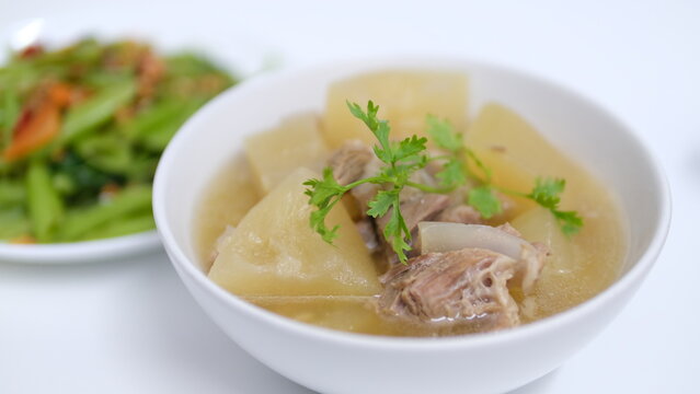  A white bowl of clear winter melon (white gourd) tender pork rib soup with blurred of stir-fried oyster sauce vegetable in the background.