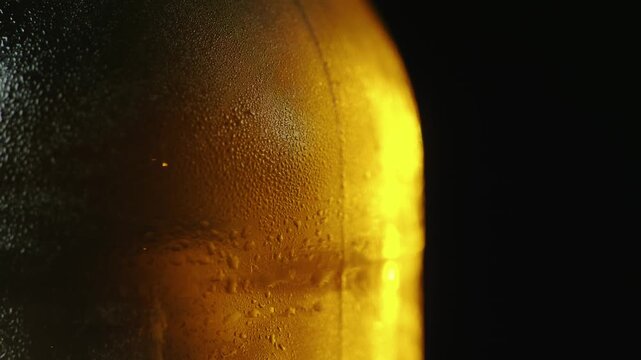 Extreme close up of cold beer bottle with water droplets on surface rotating in dark setting.