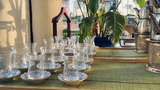 Vertical close up of glass Turkish tea cups with patterned saucers on resort buffet counter. Five stars hotel restaurant dining area with house plants background, traditional oriental tableware