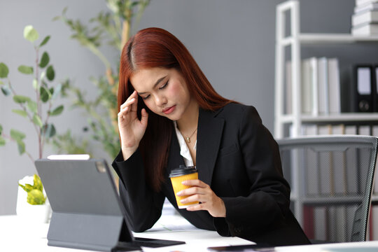 Young Asian businesswoman suffering from headache, holding coffee cup and touching her forehead while sitting at office desk and working with digital tablet.