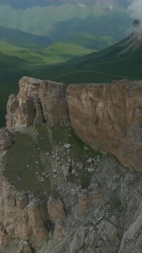 Drone view of Mount Naudzhidza with dramatic rocky cliffs, green alpine slopes and low clouds drifting over the ridge, scenic summer landscape in Russia