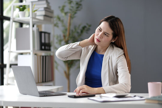 Stressed businesswoman massaging her neck, experiencing discomfort and stiffness after long hours of working on a laptop in office.