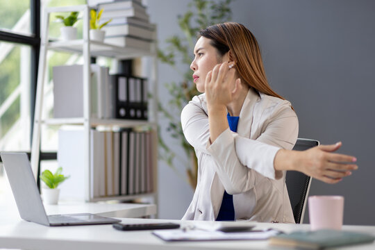 Businesswoman stretches her arms while sitting at her desk, taking a break from work. She is surrounded by office supplies and a laptop, promoting workplace wellness and productivity.