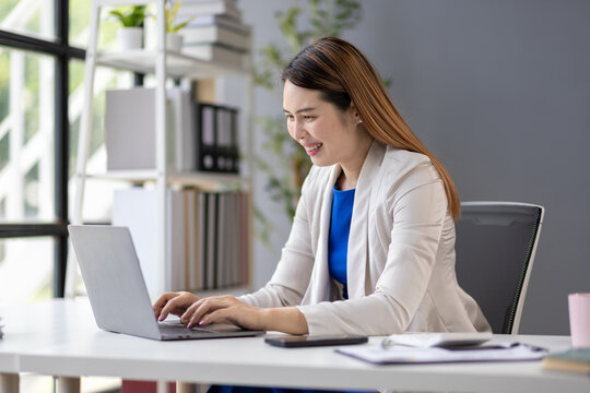 Asian businesswoman is working with laptop at office desk looking and typing on digital device screen.