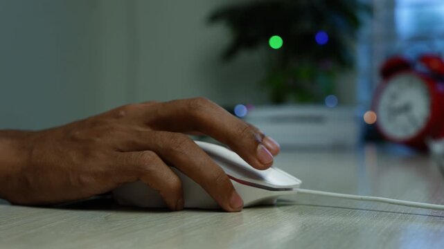 Hand using computer mouse at desk, Close-up of a hand using a white computer mouse on a desk, with a blurred red alarm clock and indoor plant in the background.