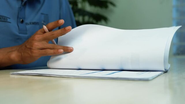 Person reviewing documents in an office, Close-up of a person in a blue uniform turning pages in a paper file on a desk, showing office paperwork, records, and document review.