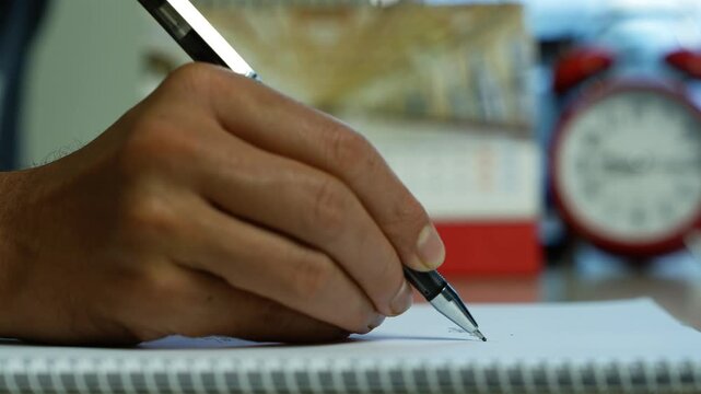 Hand writing notes in a spiral notebook, Close-up of a person writing with a pen in a spiral notebook, with a blurred alarm clock and desk items in the background.