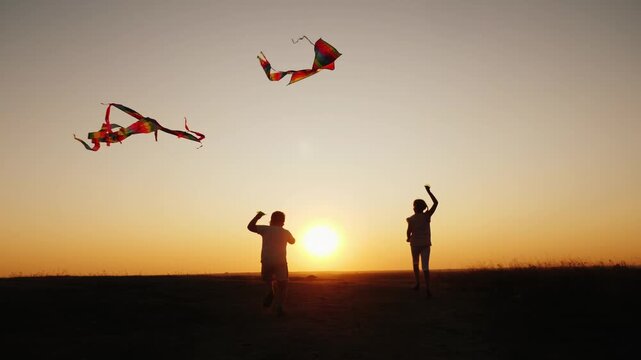 Two joyful children run across field with colorful kites during scenic golden hour sunset, cinematic slow motion video.
