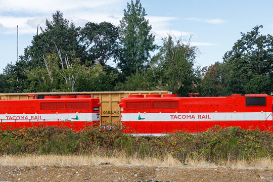 Two Tacoma Rail locomotives numbered 2200 coupled nose to nose with a tan RAILCAR spacer car, parked in tall grass along a gravel roadside.