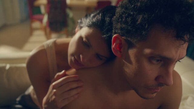 Gentle young woman leaning her head on her shirtless partners shoulder, affectionately touching his back and talking to comfort him during emotional reconciliation at home