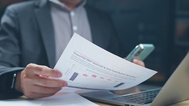 Businessman analyzing sequential ebit waterfall financial report paper holding smartphone beside laptop computer on office desk during corporate budget review meeting.