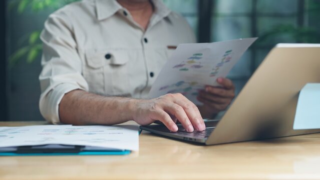 Focused engineer reviews printed process flowcharts types on laptop keyboard at wooden desk in office workspace.