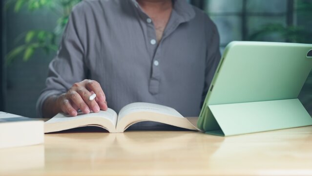 Midsection view focused adult man holding stylus pen over textbook next digital tablet on wooden table, engaged in distance learning, academic research, exam preparation, remote office work indoors.