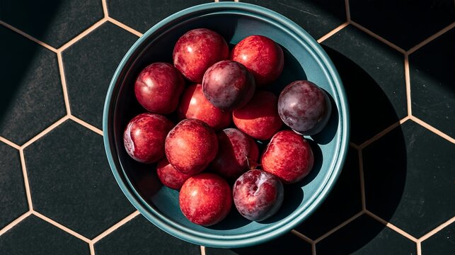 Down view of a blue bowl filled with fresh red and purple plums on a dark black hexagonal tile background, with natural sunlight shadows