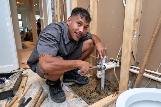 Plumber kneels, focused on pipe repair. Tools lie nearby, work area unfinished. Focused expression suggests dedication to task. Ideal for home renovation, plumbing service themes