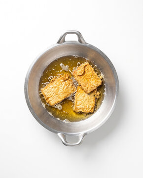 Top-down view of three pieces of battered tempeh deep-frying in hot bubbling oil inside a two-handled metal wok, isolated on a white background.