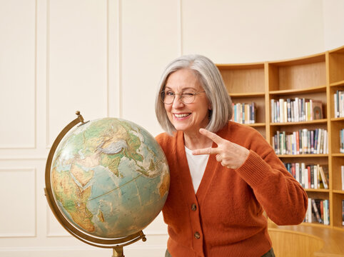 Senior woman winking and making a peace sign next to a large globe in a library, illustrating playful active aging and geography education.