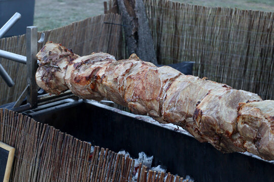 Large portion of meat turning slowly on a metal spit above a fire at an outdoor medieval fair, showing traditional roasting equipment and open air cooking methods.