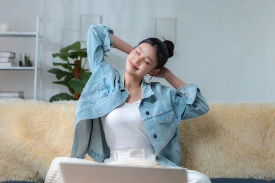 Stretching relaxation concept. Asian woman stretches on cozy sofa after laptop work at home, enjoying calm wellness break, home office, remote work, relaxation, wellness.