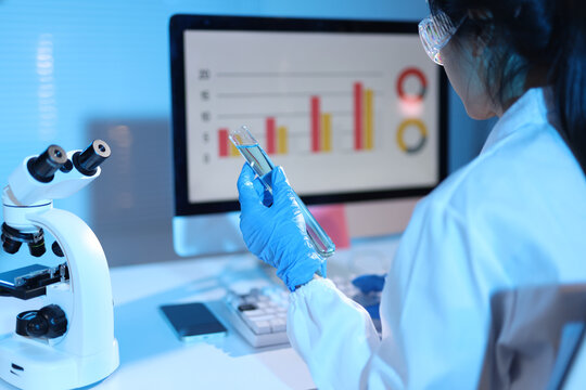 Scientist analyzing chemical liquid in laboratory with microscope. Scientist wearing protective glasses and gloves, holding a test tube with liquid, looking at a computer monitor with data charts.
