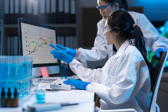 Scientists analyzing research data on computer in laboratory. Two woman scientists wearing lab coats and safety glasses analyzing experimental data on a computer screen in a dark lab.