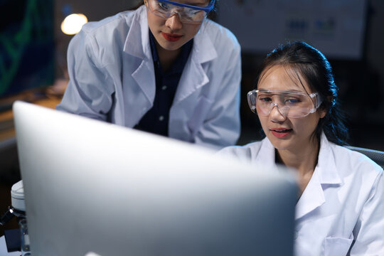 Female researchers wearing lab coats and safety glasses looking at a computer screen, analyzing data in a scientific lab.