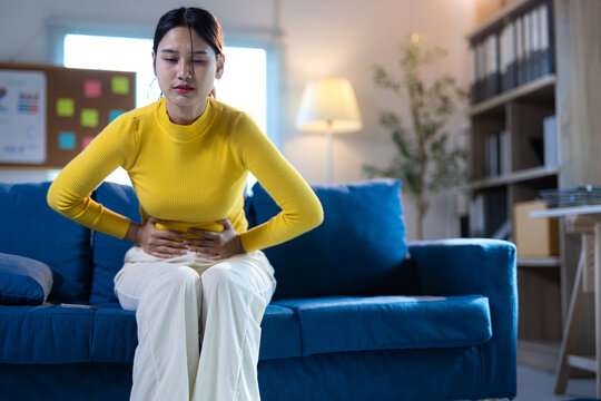 Asian woman suffering from stomach pain at home. Asian woman feeling acute abdominal pain at home, sitting on a blue sofa and holding her belly due to discomfort.