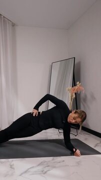 Young blonde woman in black activewear doing side plank during a home workout on a mat in a bright minimalist living room, side view