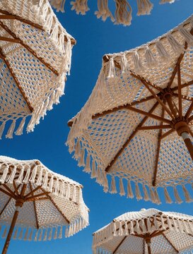 Low angle view of white macrame beach umbrellas against clear blue sky. Handmade crochet sunshades with wooden frames, long tassels decorate tropical resort. Boho style parasols provide shade for
