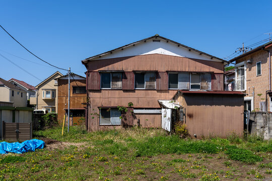 Vacant lot with weeds and an old weathered wooden house under a clear blue sky