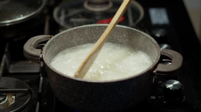 A wooden spoon stirs white coarse-ground porridge in a polka-dotted pot sitting on the stove