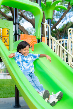 Active young Asian boy having fun sliding down a bright green slide at an outdoor playground. Healthy childhood and weekend recreation concept. Vertical Shot