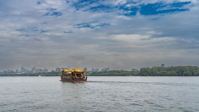 A beautiful Chinese wooden boat is floating across the picturesque city lake. Curved canopy roofs, lattice railings. Green vegetation along the shore. Multi‑story buildings against the sky. China.
 