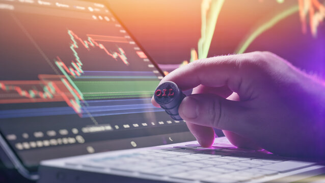 Crude oil commodity trading concept. Hand holding a miniature oil barrel in front of financial stock market charts on a laptop screen