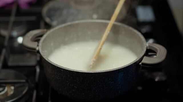 A wooden spoon stirs white coarse-ground porridge in a polka-dotted pot sitting on the stove