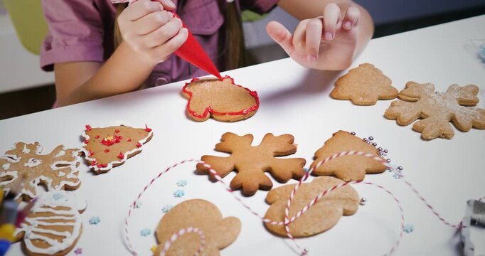 Little girl decorating gingerbread mitten with red icing at white table during christmas.