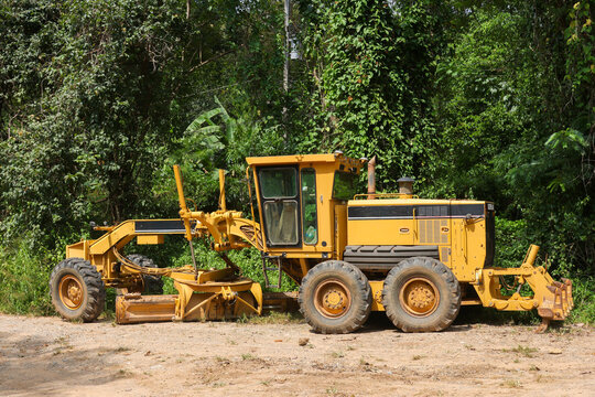 Yellow motor grader parked beside dense tropical forest with articulated blade, rugged tire, enclosed cab, and construction equipment visible in natural daylight