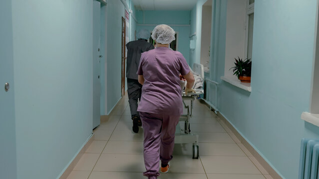 Nurse in scrubs pushing medical trolley along clean hospital corridor toward patient room, with colleague walking ahead under bright fluorescent ceiling lights