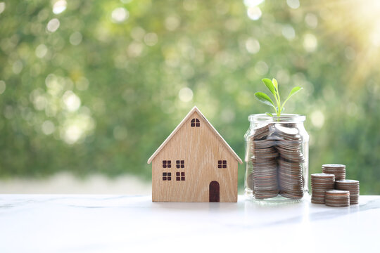 Wooden house model with coins in glass jar and growing plant on green bokeh background, concept of saving money for real estate, property investment and eco-friendly home planning.