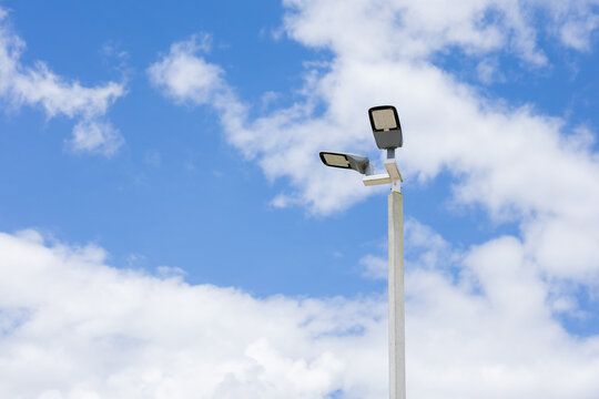 Double floodlight pole against blue sky with white cloud, outdoor lighting fixture creating clean low angle composition for infrastructure and energy themes