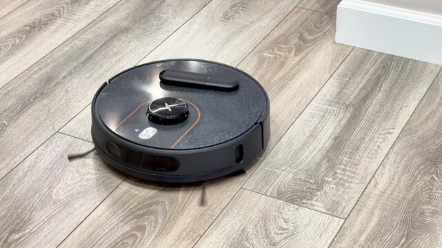 Modern black robotic vacuum cleaner cleaning a light wooden laminate floor indoors. Smart wireless home appliance sweeping dust in a cozy living room or apartment house. Domestic automatic hygiene. 