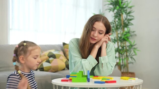 Mother watches and supports her young daughter playing with colorful building blocks at home, enjoying creative play, learning, parenting, family time together.