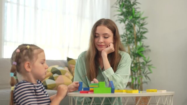 Young daughter builds colorful blocks while mother watches with interest at home, sharing creative play, learning activity, quality time, family bonding together.