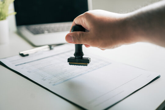 Rubber stamp approving financial document on office desk, businessman signing contract for business agreement and legal approval process with laptop in background