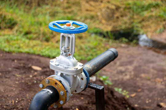 Outdoor industrial water valve with blue handwheel and pipe connection beside shallow stream and grassy soil, showing utility infrastructure, metal hardware, maintenance access, and environmental