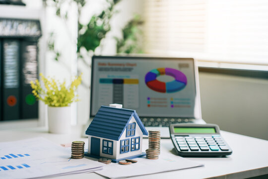 Real estate investment planning with miniature house model and coin stacks on financial data charts at office desk for residential property purchase concept