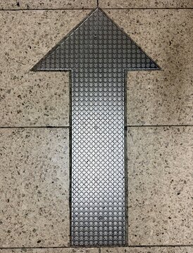 Overhead view of a textured metal arrow embedded in a speckled tile floor, pointing straight ahead.

