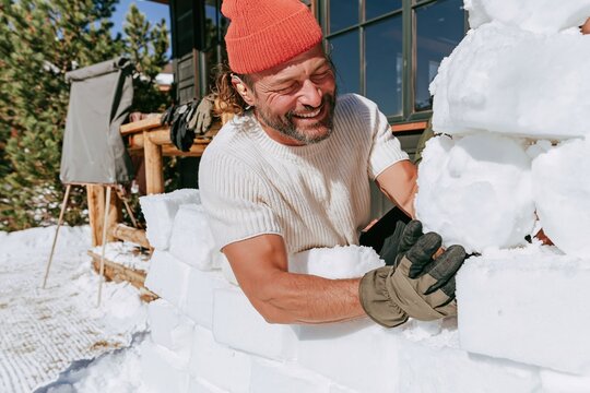 Snow wall being built by a grinning man in a red beanie, busily packing cold white blocks on a lodge terrace for team building and fun