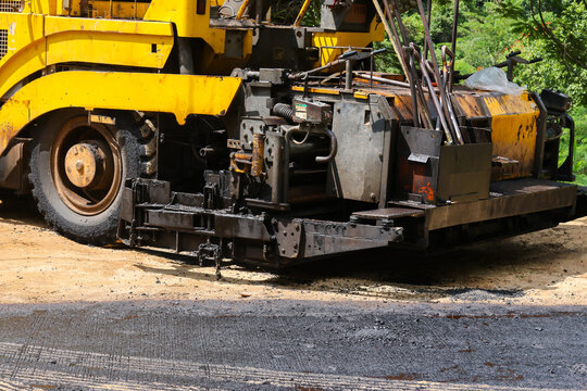 Yellow asphalt paver and roller prepare fresh road surface at construction site, showing heavy machinery, dark aggregate, dusty ground, and rugged equipment in operation